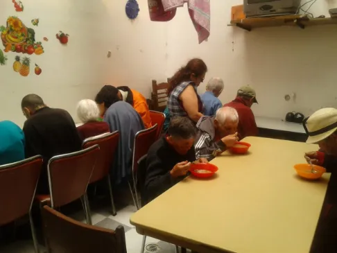 Personas mayores sentadas a una mesa larga comiendo sopa en un comedor comunitario