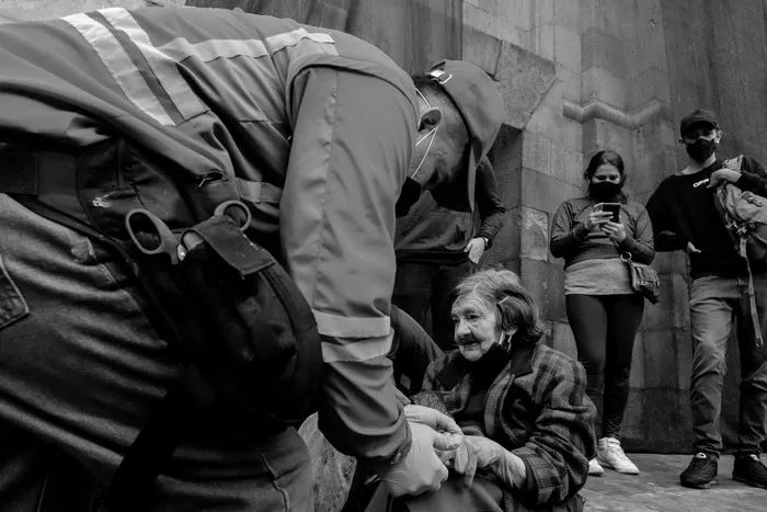Un agente inclinado sobre una persona mayor en el suelo, con manifestantes con tapabocas alrededor