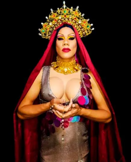 Artista drag con corona dorada, manto rojo y joyas doradas sobre fondo negro