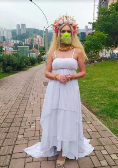Artista drag con vestido blanco, corona de flores y tapabocas camina por un sendero de un parque