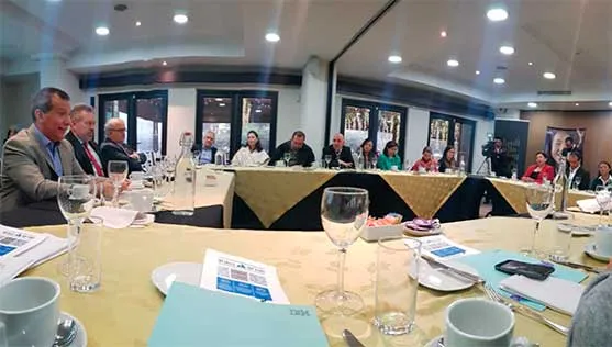 Asistentes sentados en mesas redondas con copas y carpetas durante la presentación del acuerdo