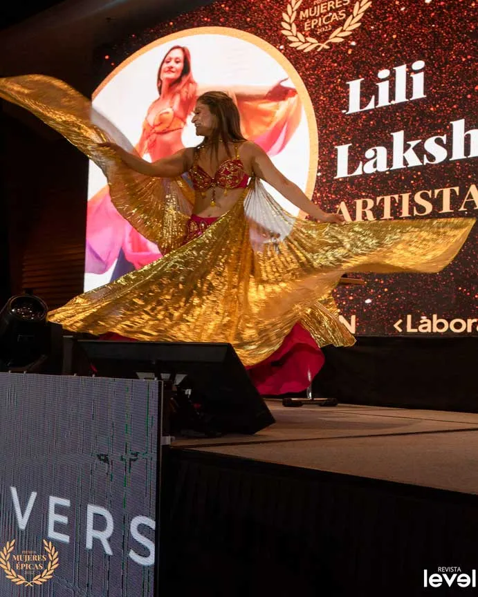 Bailarina con alas doradas en el escenario, junto a la pantalla «Lili Laksh, artista», en Mujeres Épicas de Revista Level