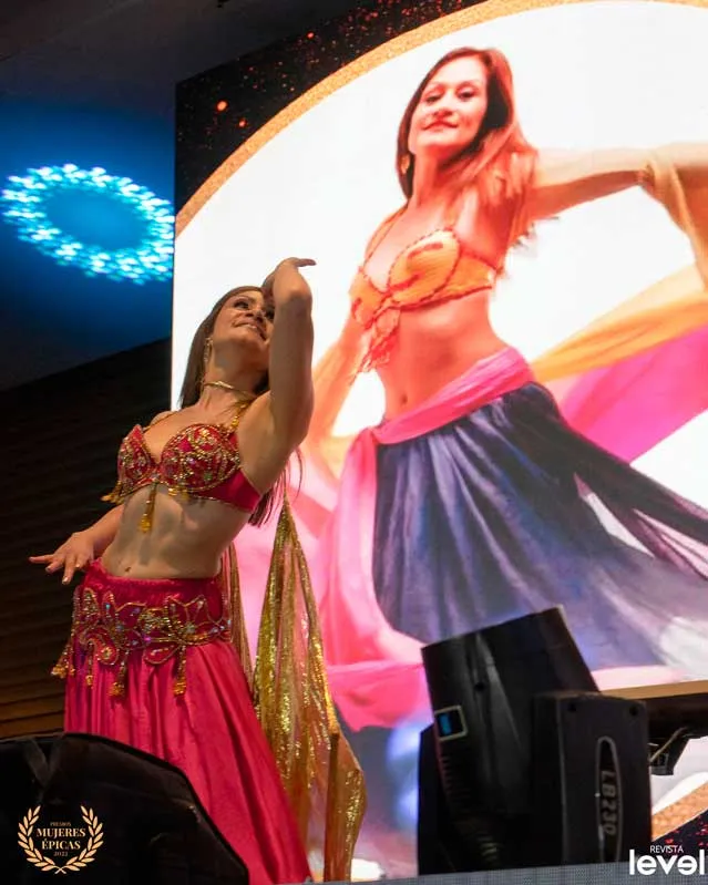 Bailarina de danza del vientre con traje rosa y dorado junto a una pantalla, en Mujeres Épicas de Revista Level