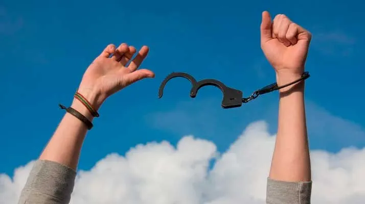 Brazos alzados contra un cielo azul con nubes, una muñeca con esposas abiertas que simbolizan la libertad