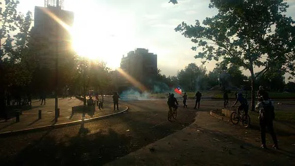 Calle con humo al atardecer, con el sol entre los árboles, personas y ciclistas durante una protesta