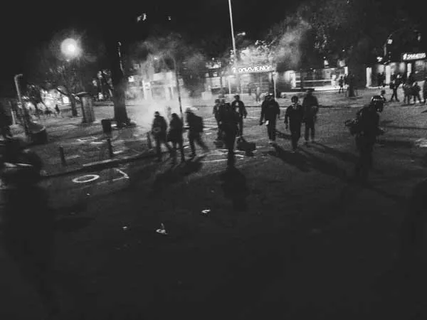 Calle nocturna con humo y personas dispersas durante una protesta, en blanco y negro
