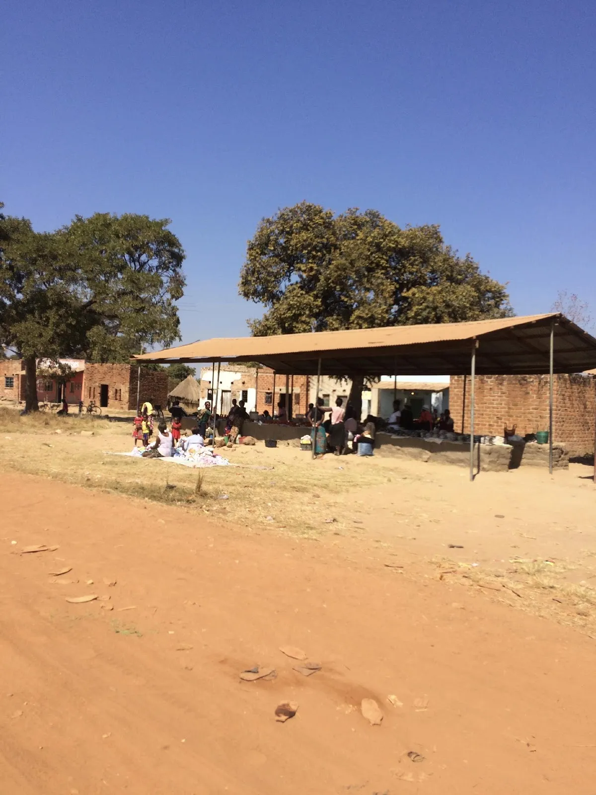 Camino de tierra roja hacia un cobertizo con techo bajo el que hay personas, en Zambia
