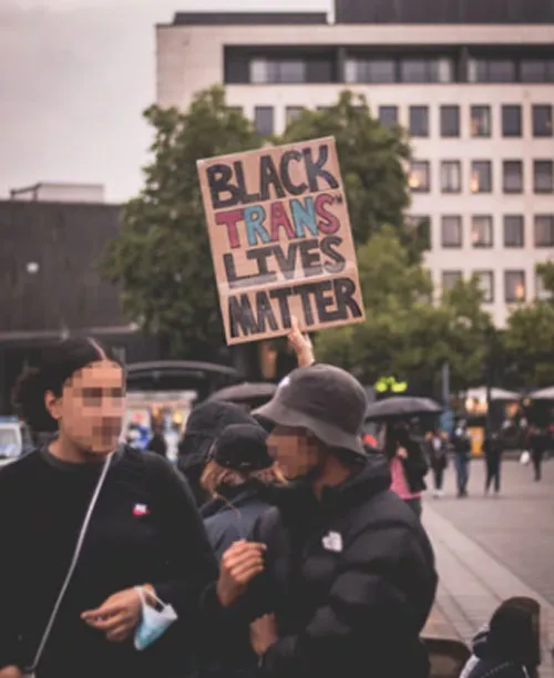 Dos personas con abrigo en una plaza, tras ellas un cartel «Black Trans Lives Matter»