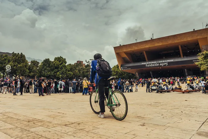 Ciclista cruza una plaza frente al edificio de Fundación EPM, con una multitud reunida al fondo