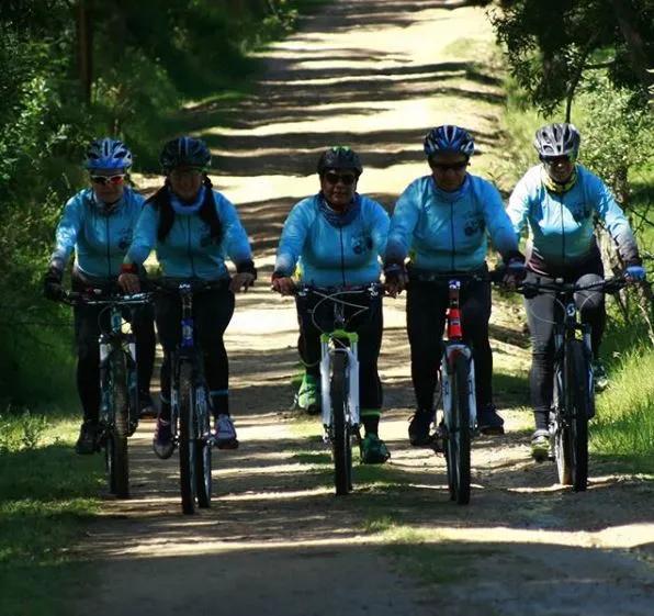 Cinco ciclistas con uniforme azul turquesa y casco pedalean por un camino de tierra entre árboles