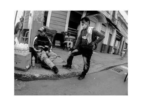 Dos hombres con tapabocas conversan en una esquina de la ciudad, uno sentado y otro de pie, en blanco y negro