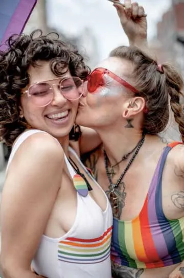 Dos mujeres con gafas de sol y tops con los colores del arcoíris se besan en la mejilla y sonríen durante una celebración