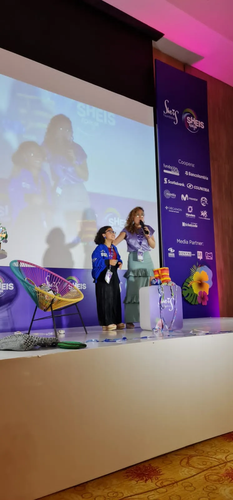 Dos mujeres conversan de pie en el escenario del She is Global Forum ante una pantalla con imágenes