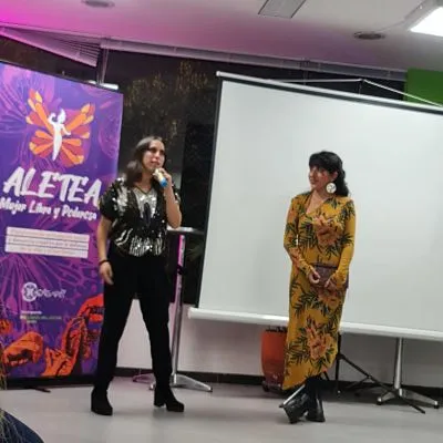 Dos mujeres hablan en un escenario junto al pendón «Aletea, Mujer Libre y Poderosa» y una pantalla de proyección