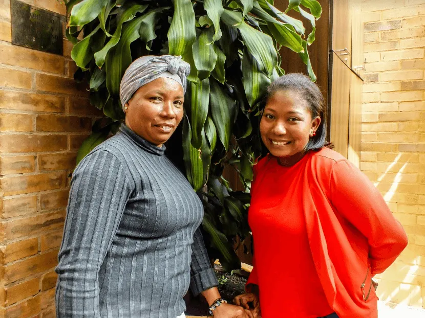 Dos mujeres posan sonriendo junto a un muro de ladrillo y plantas, una con pañuelo gris y otra con blusa roja