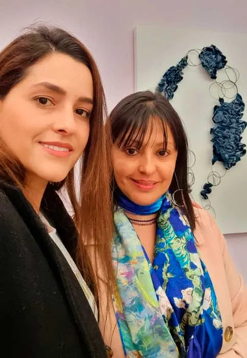 Dos mujeres sonrientes en un selfi frente a una obra de joyería azul colgada en la pared