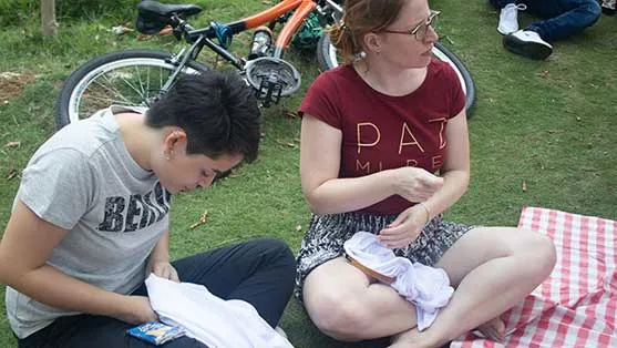 Dos personas bordando telas blancas sentadas en el pasto junto a una bicicleta