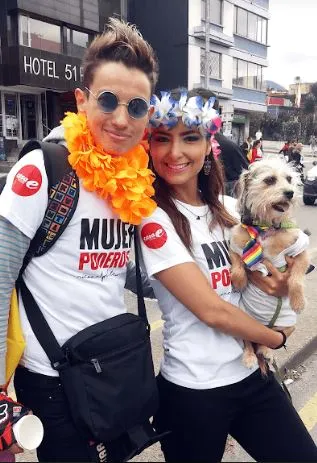 Dos personas con camiseta blanca «Mujer poderosa» se abrazan con un perro pequeño en brazos
