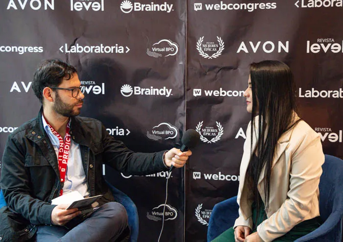 Un hombre entrevista con un micrófono a una mujer sentada frente al panel de Revista Level, Braindy y Avon