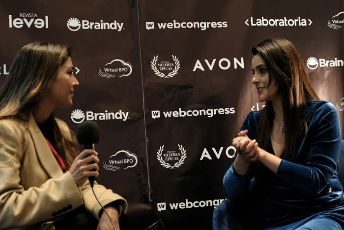 Una mujer entrevista con un micrófono a otra mujer frente al panel de Revista Level, Braindy y Avon