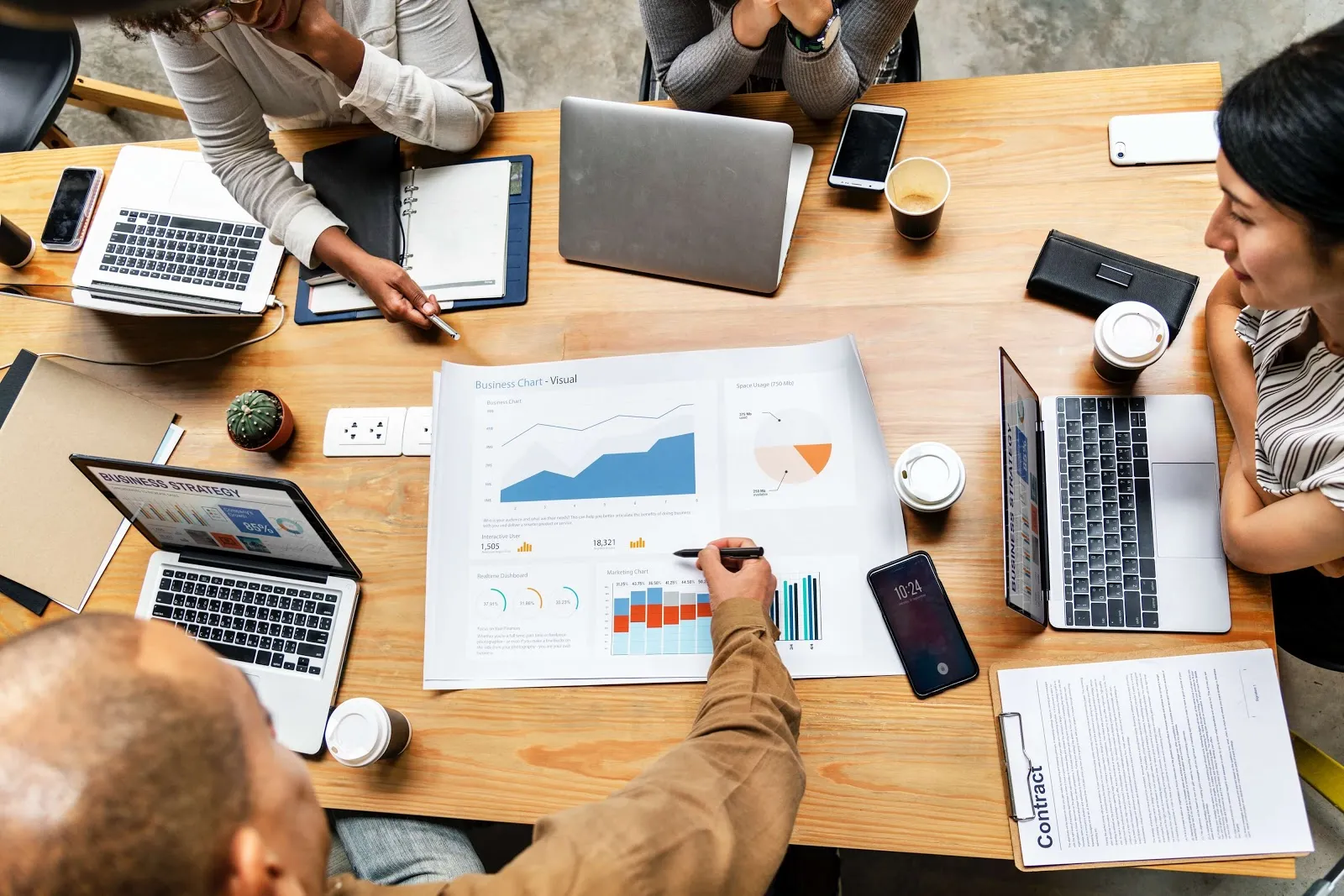 Equipo de trabajo reunido alrededor de una mesa de madera con portátiles, celulares y un informe con gráficos