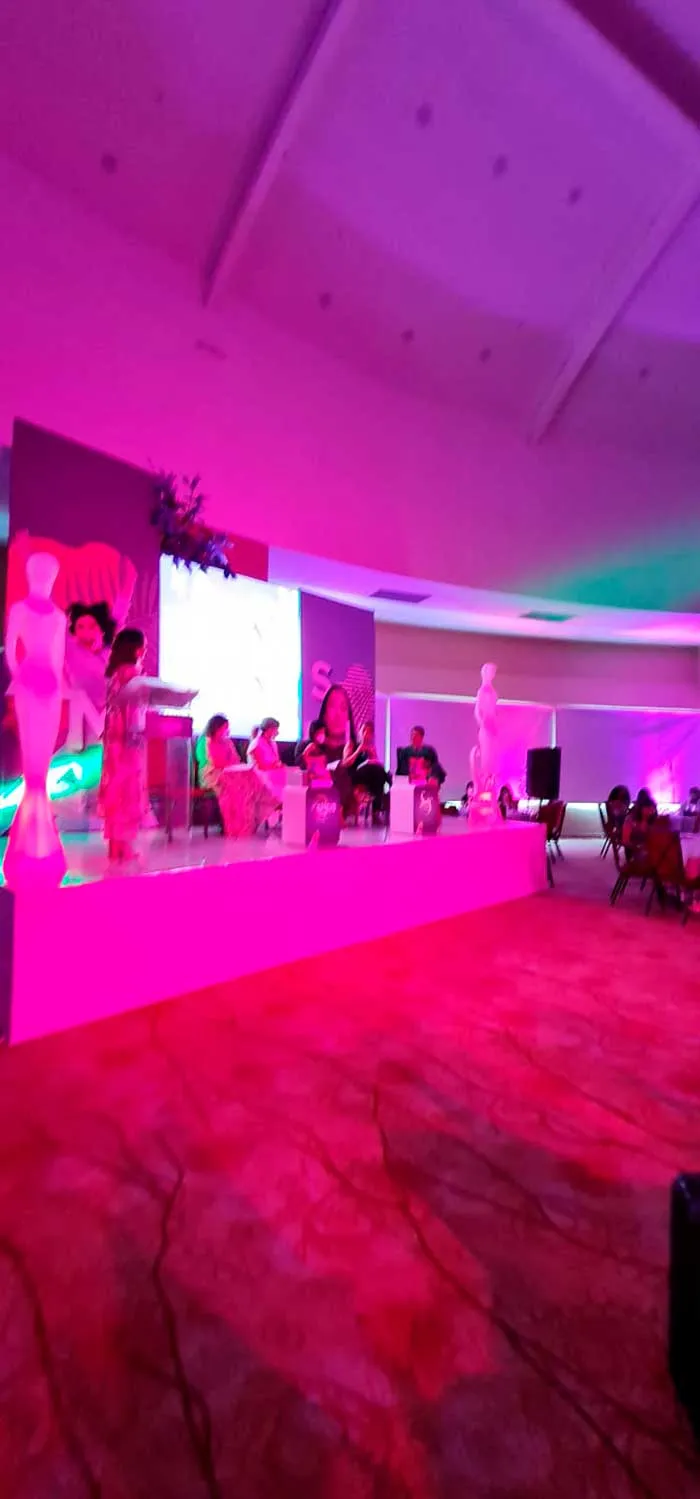Escenario del She is Global Forum iluminado en rosa, con oradoras y asistentes al fondo