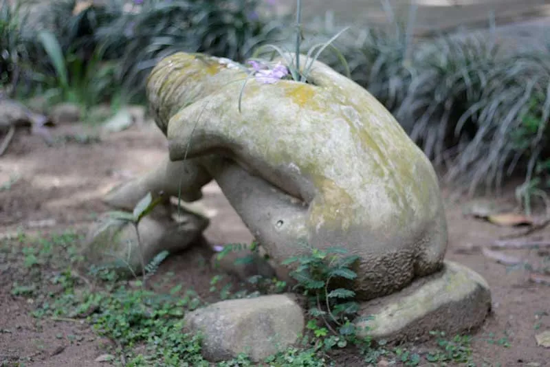 Escultura de una figura encogida cubierta de musgo y plantas en un jardín