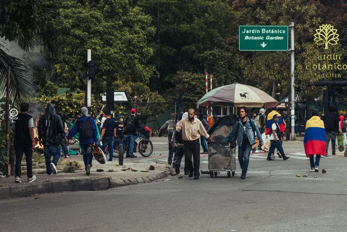 Manifestantes y un carrito callejero en una esquina, con una persona envuelta en la bandera de Colombia