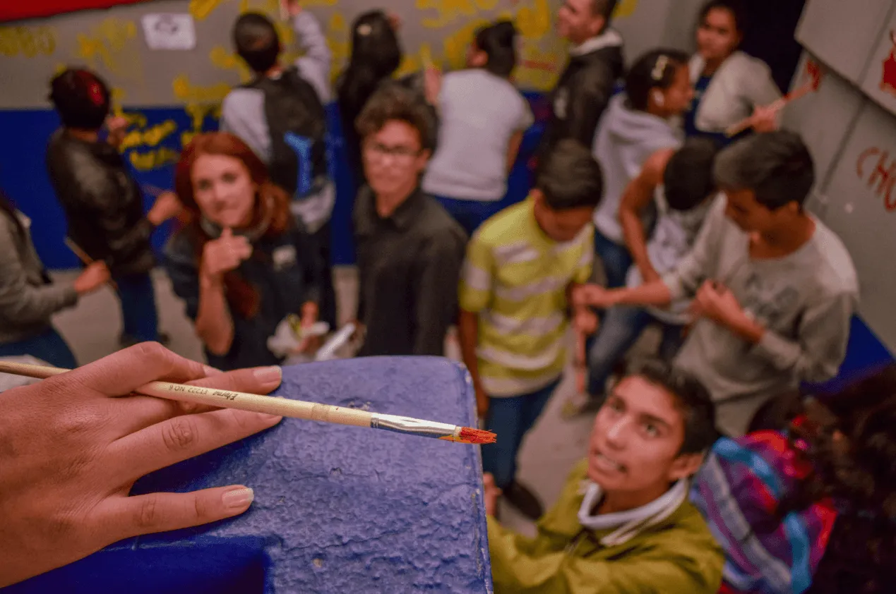 Estudiantes reunidos en un patio alrededor de una pared que pintan de azul, con un pincel en primer plano
