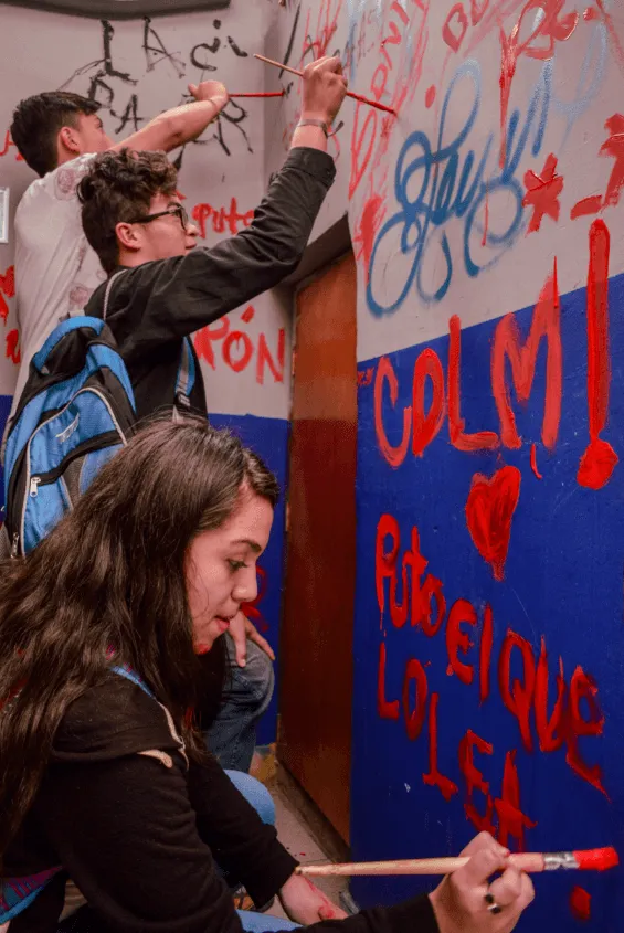 Estudiantes pintan con brochas un mural azul y rojo con corazones y frases en la pared