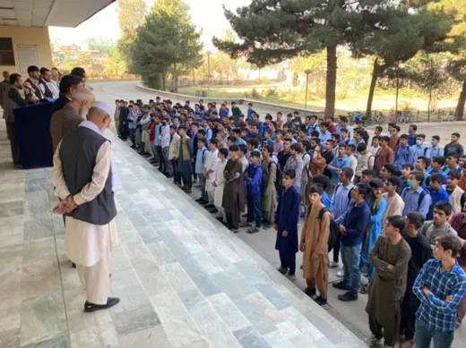 Cientos de estudiantes varones formados en fila frente a un edificio escolar en Kabul, con un hombre mayor de pie al frente