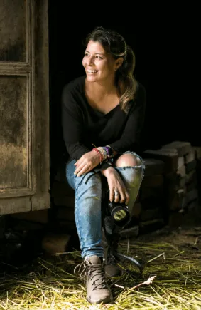 Fotógrafa sonriente sentada en un banco dentro de un granero, con jeans rotos y botas