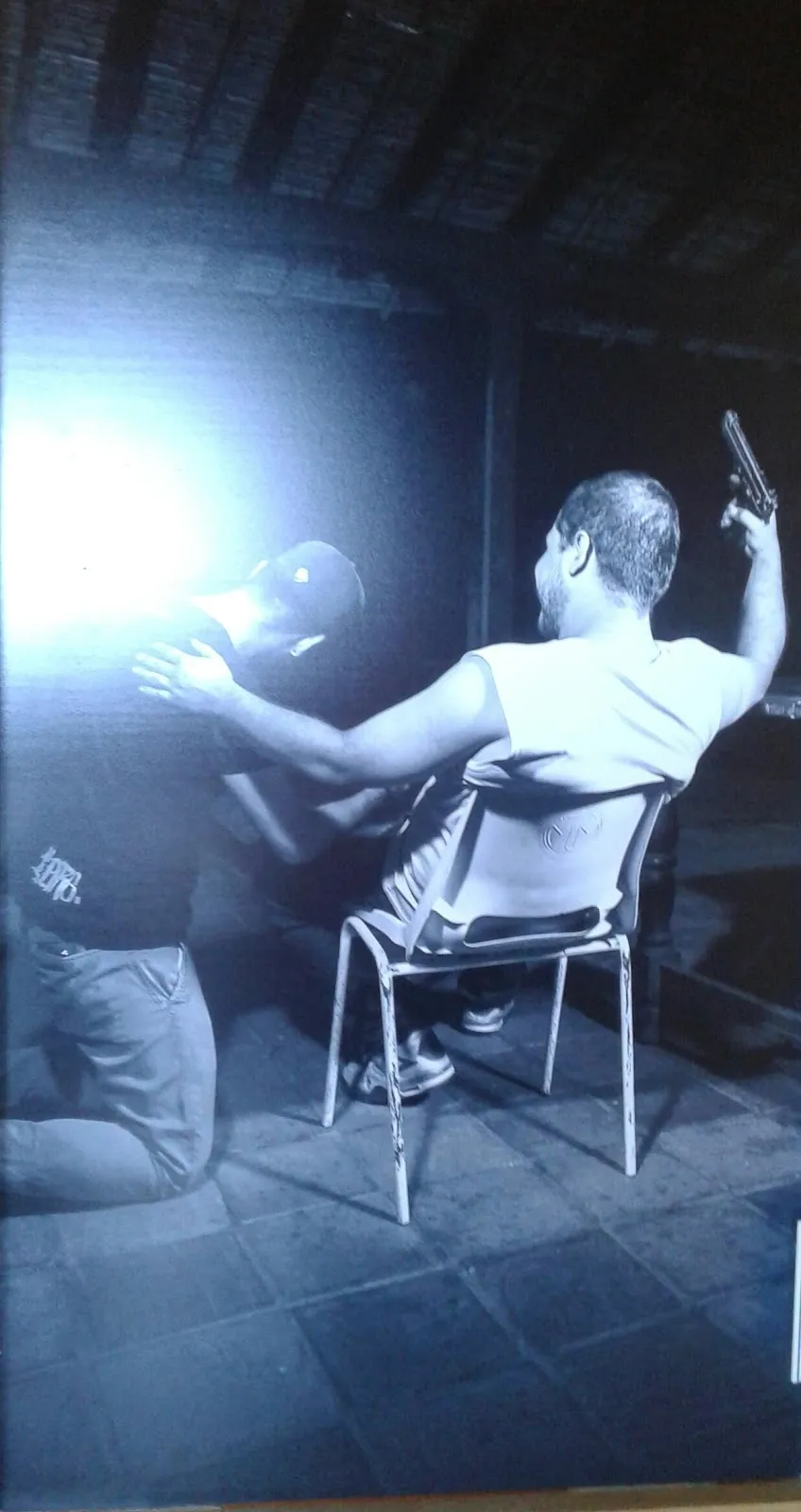 Hombre sentado en una silla apuntado con un arma por una persona con una luz intensa detrás, de la serie «¡Juguemos!»