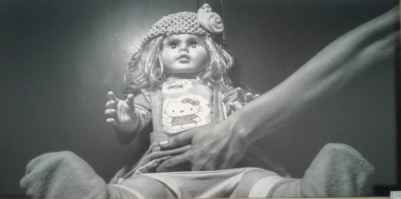 Muñeca sentada entre las piernas de una persona mientras unas manos la rodean, imagen en blanco y negro sobre el abuso infantil
