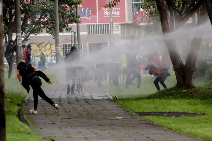 Chorro de un carro hidrante sobre manifestantes que corren en un parque