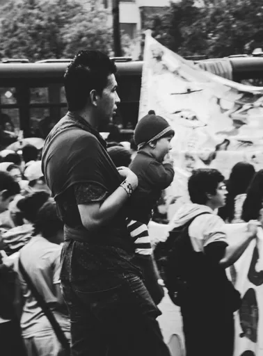 Hombre carga a un niño con gorro entre la multitud de una marcha, junto a una pancarta