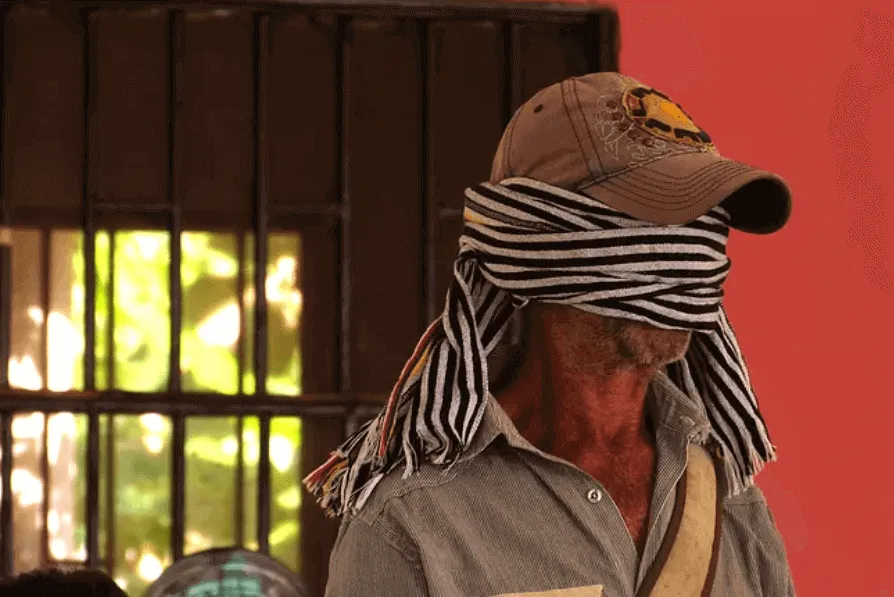 Hombre con gorra y un pañuelo a rayas sobre los ojos, con la cabeza inclinada, frente a un muro rojo