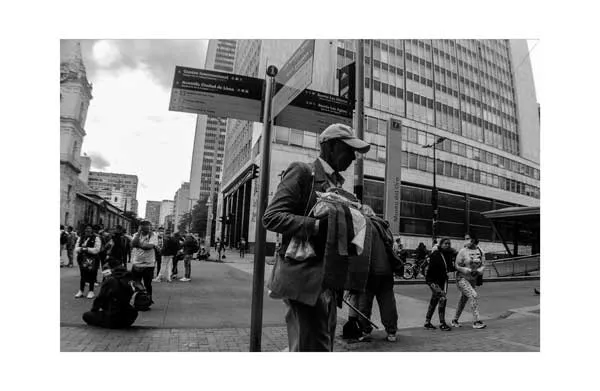 Hombre con gorra y tapabocas junto a un poste de señalización en una calle con edificios altos, en blanco y negro