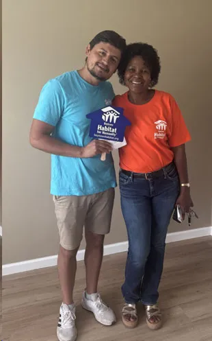 Un hombre y una mujer con camisetas de Habitat for Humanity posan dentro de una casa, él con un cartel azul
