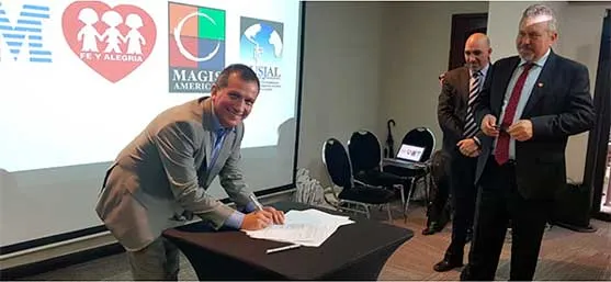 Dos hombres firman un documento en una mesa mientras otros observan de pie, ante una pantalla con logos