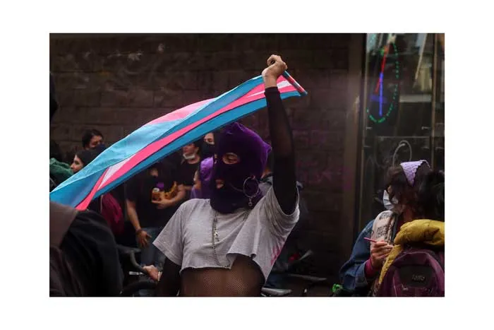 Manifestante con pasamontañas morado levanta una bandera trans en el 8 de marzo de 2021