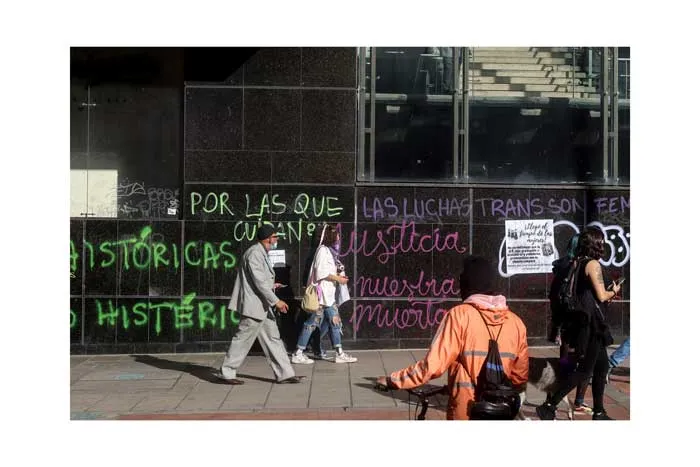 Personas caminan frente a un muro con pintadas verdes y moradas sobre las luchas trans y feministas