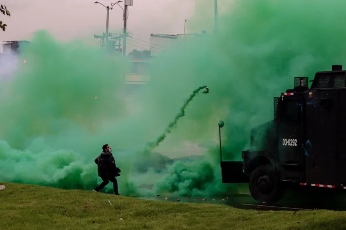 Humo verde de gas lacrimógeno junto a un vehículo blindado de la policía y una persona corriendo en una protesta en Colombia