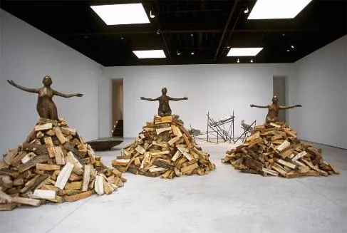 Instalación de Kiki Smith (2002) con tres figuras femeninas de brazos abiertos sobre pilas de leña en una sala de museo
