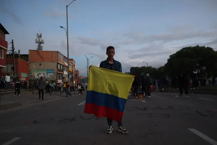 Joven de pie sostiene una bandera de Colombia en una calle al atardecer, con manifestantes al fondo