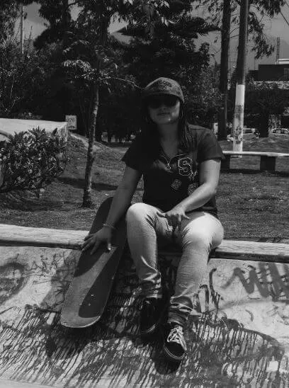 Joven con gorra sentada en el borde de una rampa de skatepark con su patineta al lado, en blanco y negro