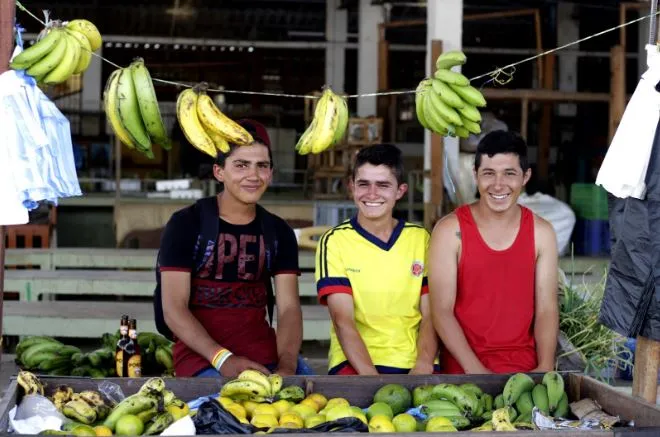Tres jóvenes sonrientes detrás de un puesto de frutas con racimos de plátano colgados