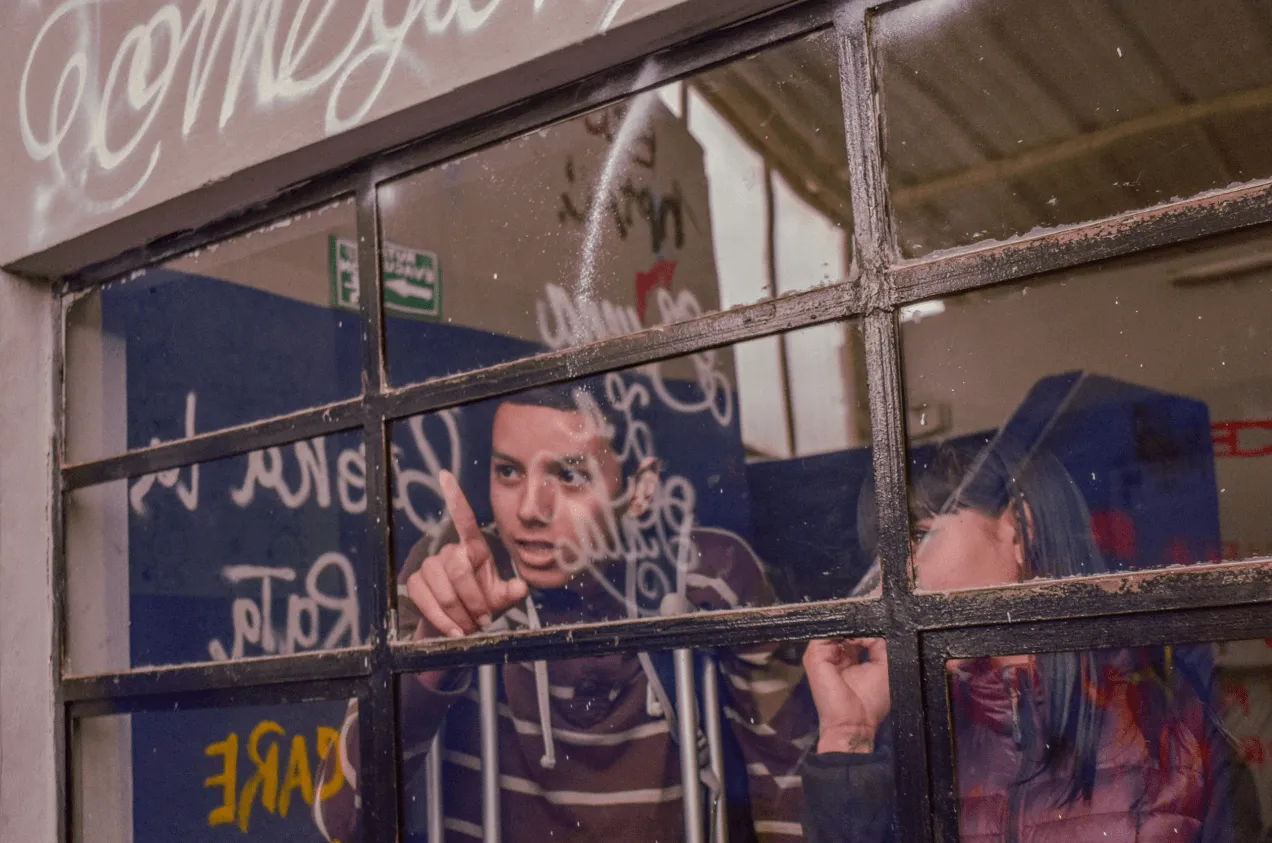 Jóvenes tras una ventana con vidrios rayados observan el mural pintado en el exterior