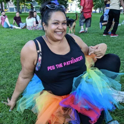 Luna Matatas sonríe sentada en el césped con una camiseta «Peg the patriarchy» y una falda de tul de colores del arcoíris