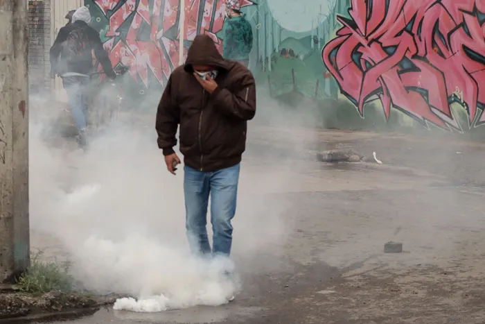 Manifestante con capucha camina entre humo de gas lacrimógeno frente a un muro con grafitis